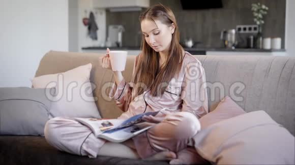 美丽的黑发女孩穿着丝绸睡衣晚上坐在客厅的沙发上喝茶看书视频的预览图