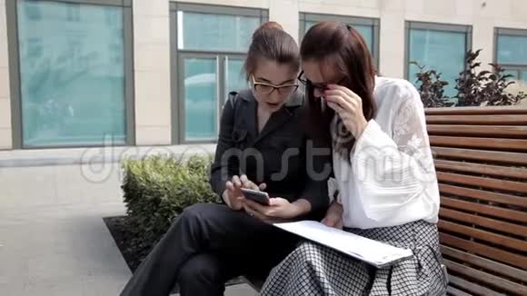 两个年轻的商业女孩坐在商业中心附近的公园里笑着看着电话里的商业想法视频的预览图