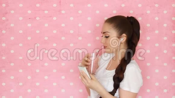 美丽的快乐女孩戴着耳机喝水果或浆果冰沙年轻女子拿着粉红色的鸡尾酒视频的预览图