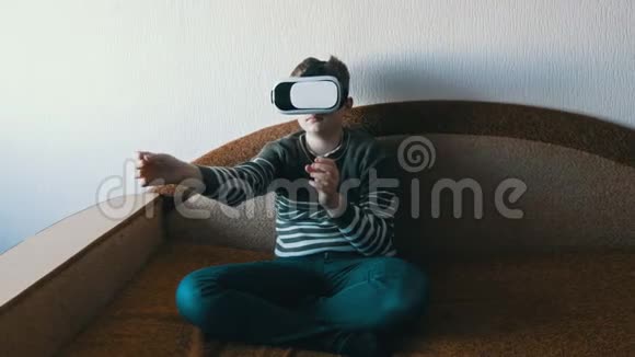 戴着虚拟现实的白色眼镜或虚拟现实的青少年男孩头上坐在房间里的沙发上玩耍视频的预览图