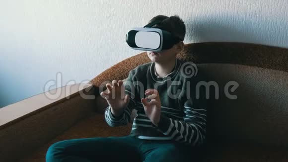 戴着虚拟现实的白色眼镜或虚拟现实的青少年男孩头上坐在房间里的沙发上玩耍视频的预览图