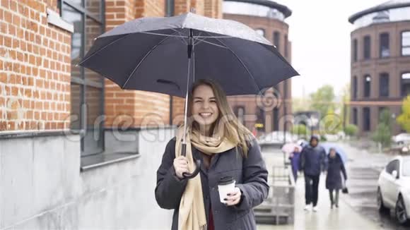 一个开朗的金发女商人带着咖啡和雨伞在街上视频的预览图