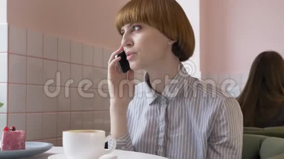 一个年轻漂亮的红发女孩坐在咖啡馆里用智能手机接电话打电话视频的预览图