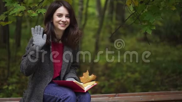 一个笑容满面的黑发女人手里拿着一本书在秋天的公园里挥手视频的预览图