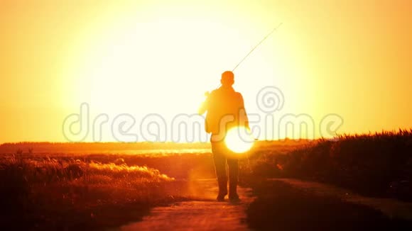 渔夫在日落的天空背景下散步那个人拿着一根鱼竿和钓具从远处下去视频的预览图