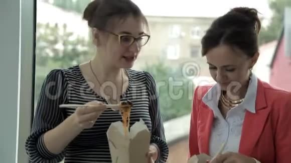 两个年轻的商业女孩在银行工作在窗口吃午餐吃中国面条讨论生意视频的预览图