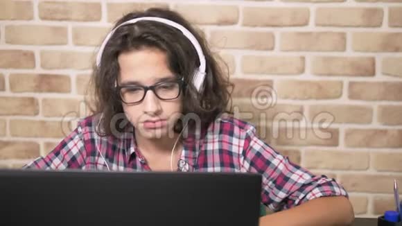酷酷的少年男孩一头卷曲的黑发穿着格子衬衫戴着耳机跳舞坐在笔记本电脑前视频的预览图