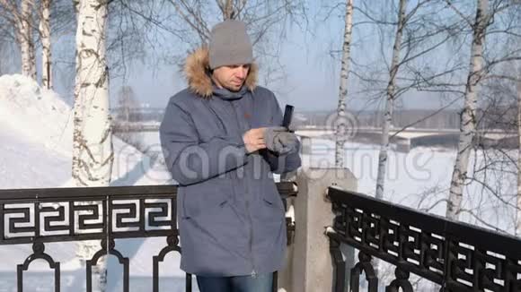 穿着蓝色夹克带着毛皮罩的人在冬季公园用手套擦拭手机屏幕视频的预览图