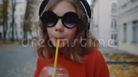 特写镜头漂亮的时髦少年用耳机听音乐用塑料杯喝奶昔微笑着走在街上视频的预览图