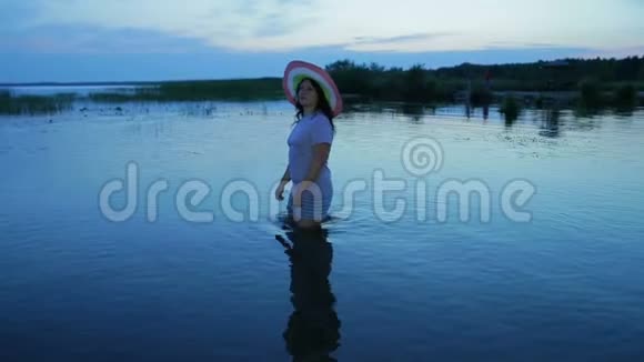一个穿着短裤和帽子的年轻女孩沿着湖边散步欣赏日落视频的预览图