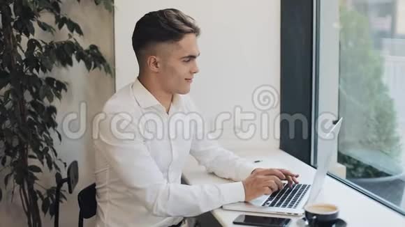 快乐的年轻商人在窗户附近的咖啡馆里用笔记本电脑工作商业事业成功自由职业者求职视频的预览图