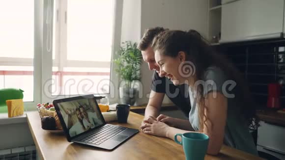 年轻漂亮的白种人夫妇坐在笔记本电脑前的桌子上和他们快乐的积极的晒黑的朋友一起飞翔视频的预览图