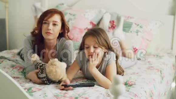 快乐的母亲和可爱的女儿在电视上看卡通电影一边用遥控器和微笑躺在床上视频的预览图