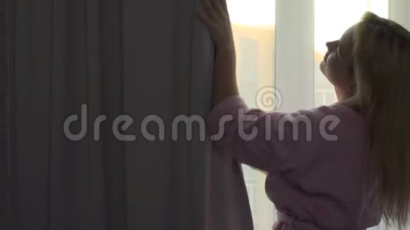 一位穿着家庭浴袍的年轻女子早上醒来时拉开窗帘视频的预览图