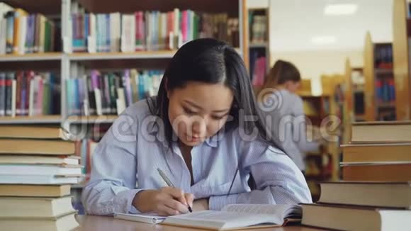 年轻漂亮的亚洲女学生坐在桌子旁周围是大图书馆里成堆的书从大图书馆改写课文视频的预览图