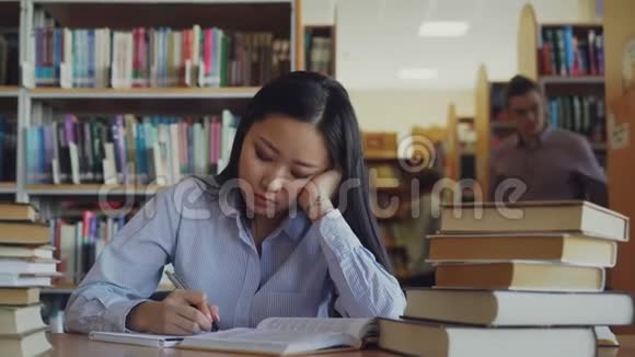 年轻的亚洲少女正坐在大图书馆的书桌前重写书中的文字她又累又累看上去视频的预览图