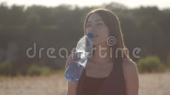 在炎热的夏日里口渴的年轻女子在日落时喝着塑料瓶里的水视频的预览图
