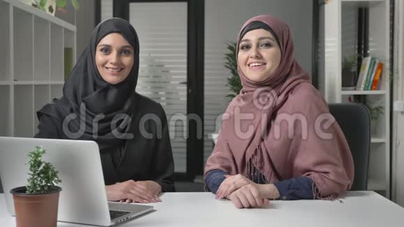 两个戴头巾的年轻女孩坐在办公室里看着摄像机微笑积极60帧秒视频的预览图