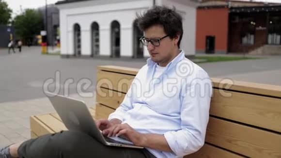 戴眼镜的年轻商人坐在城市的长凳上打字视频的预览图