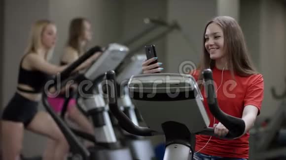 穿红色衬衫的漂亮女孩在健身车上大力工作并在新健身房与手机交谈视频的预览图