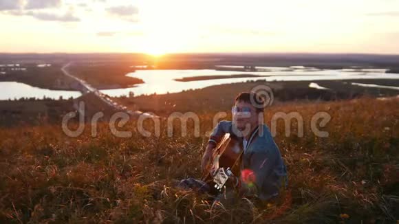 戴着圆眼镜的男孩在日落时分的田野里弹吉他仰望天空视频的预览图