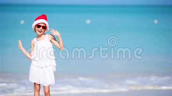 圣诞节假期戴着圣诞帽的小可爱女孩在白色海滩上开心微笑的孩子看着摄像机视频的预览图