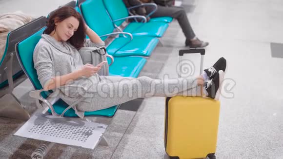 疲惫的女人正坐在机场候机大厅的座位上休息双腿搭在黄色的手提箱上看着磁带新闻视频的预览图