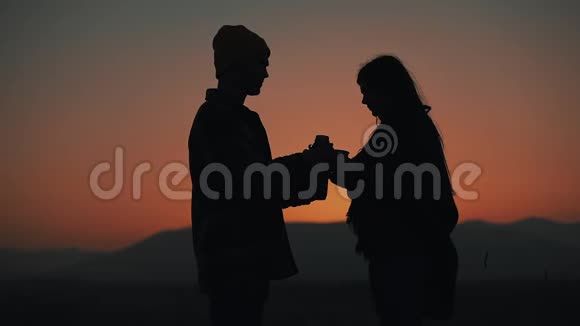 在美丽的夕阳背景下一对相爱的年轻夫妇用热水瓶喝茶的剪影视频的预览图