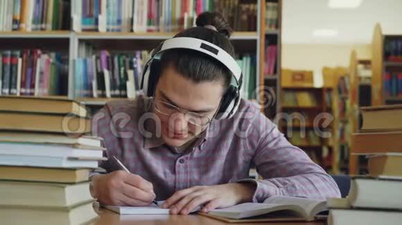 年轻的白种人男学生带着大耳机坐在图书馆的桌子旁他身边有很多书他是视频的预览图