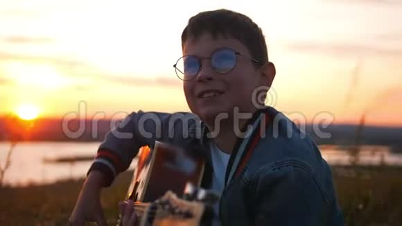 戴着圆眼镜的男孩在日落时分的田野里弹吉他仰望天空视频的预览图