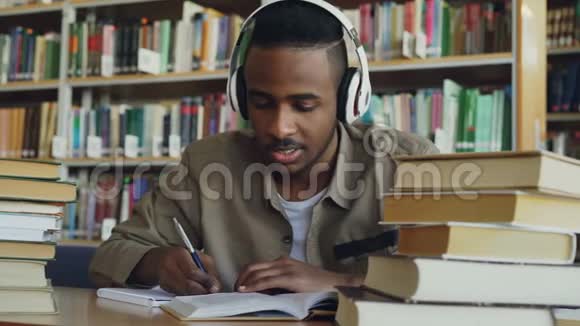 英俊的非裔美国男学生戴着耳机坐在宽敞的图书馆里的桌子旁听音乐视频的预览图