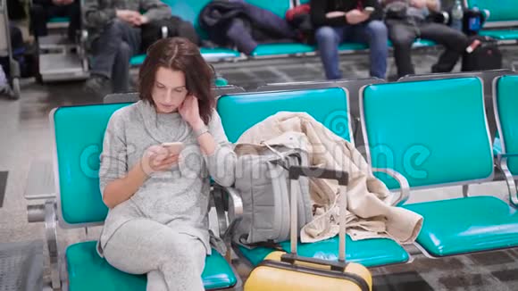 在飞机起飞前独自一人的年轻女子坐在机场候机室的椅子上在她的手机着留言视频的预览图