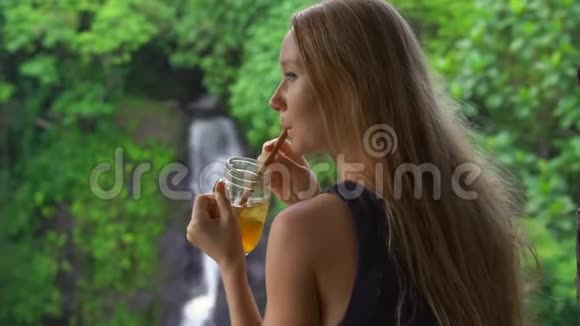一位年轻的女士在一家美丽的咖啡馆里在峡谷和瀑布上有着史诗般的景色喝着冰茶前往巴厘岛视频的预览图
