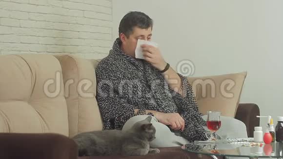 一个有流感迹象的人用毯子盖着坐在沙发上的公寓里把鼻涕吹成纸视频的预览图