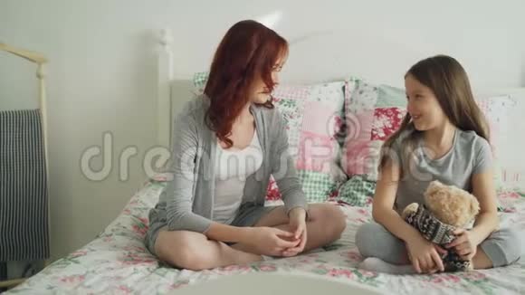 年轻细心的母亲和开朗的小女儿在卧室里坐在床上谈论和分享秘密视频的预览图