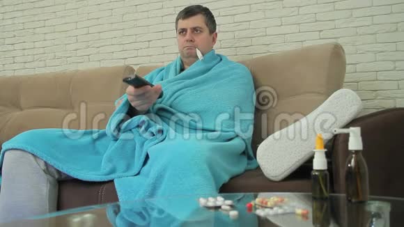 一位有感冒症状的成年男性坐在沙发上的毯子里测量体温并打开电视视频的预览图