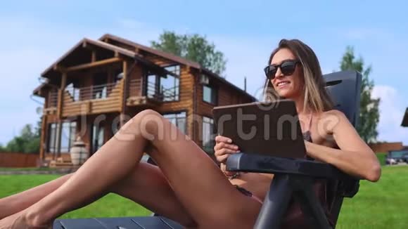 美丽苗条的女人躺在阳光下的休息室里穿着泳衣在一所大乡村房子的背景上放着一块平板电脑视频的预览图