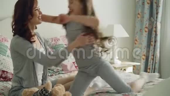 快乐的小女儿在舒适的床上坐着陪着年轻快乐的妈妈玩玩具熊视频的预览图