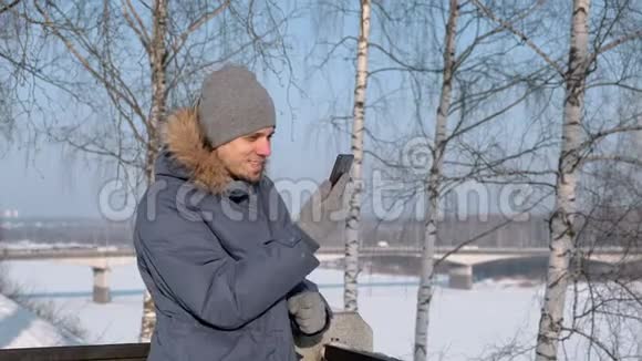 穿着蓝色羽绒服带皮帽的人在冬季公园里用手机视频聊天视频的预览图