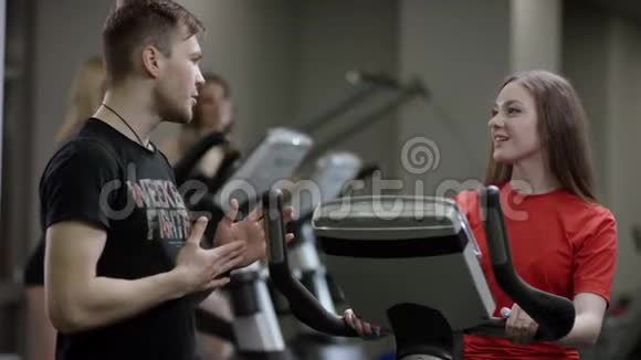 可爱的女孩穿着红色衬衫骑着运动自行车教练来了在新的健身房里贪婪地对待她视频的预览图