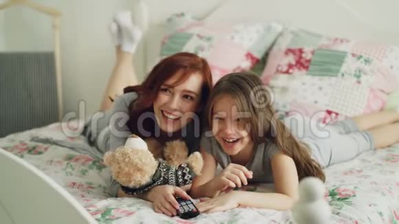 早上躺在家里的时候开心的妈妈和可爱的女儿在电视上看有趣的卡通电影视频的预览图