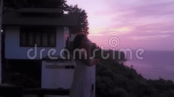 年轻女子站在阳台上看着夕阳喝着热咖啡上面有美丽的山景视频的预览图