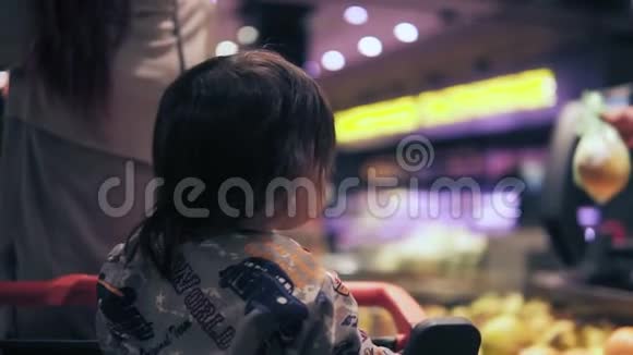一个蹒跚学步的孩子坐在购物车和女顾客采摘橙色包裹的背面镜头视频的预览图