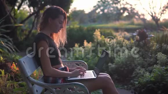 学生女孩在户外学习年轻的金发女郎穿着黑色上衣和短裤坐在公园的长凳上在笔记本电脑上工作美丽的自然视频的预览图