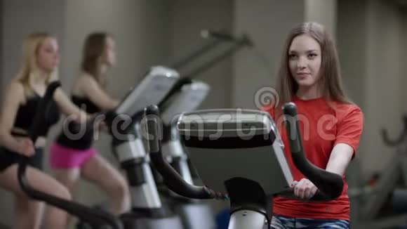 可爱的女孩穿着红色衬衫大力工作在运动自行车只是在新的健身房工作的背景下视频的预览图