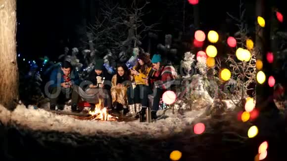 冬天森林里的年轻人坐在火边坐在电话里的人视频的预览图