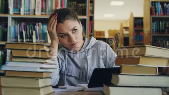 美丽的白种人少女坐在大学图书馆的桌子上手里拿着数字平板电脑她很累而且视频的预览图
