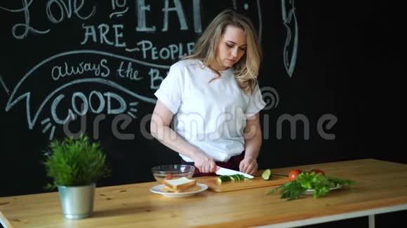 美丽的年轻女子用菜刀在木切割板上的圆片上切绿黄瓜准备视频的预览图