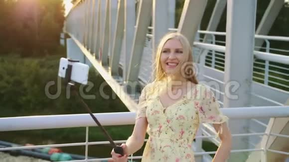 女人在桥上自拍站在人行桥上的一位开朗漂亮的金发女郎和monopod一起自拍视频的预览图