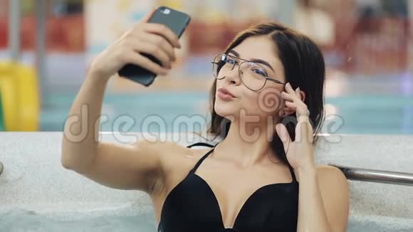 漂亮的黑衣年轻女子在智能手机上用全新的按摩浴缸或游泳池自拍视频的预览图